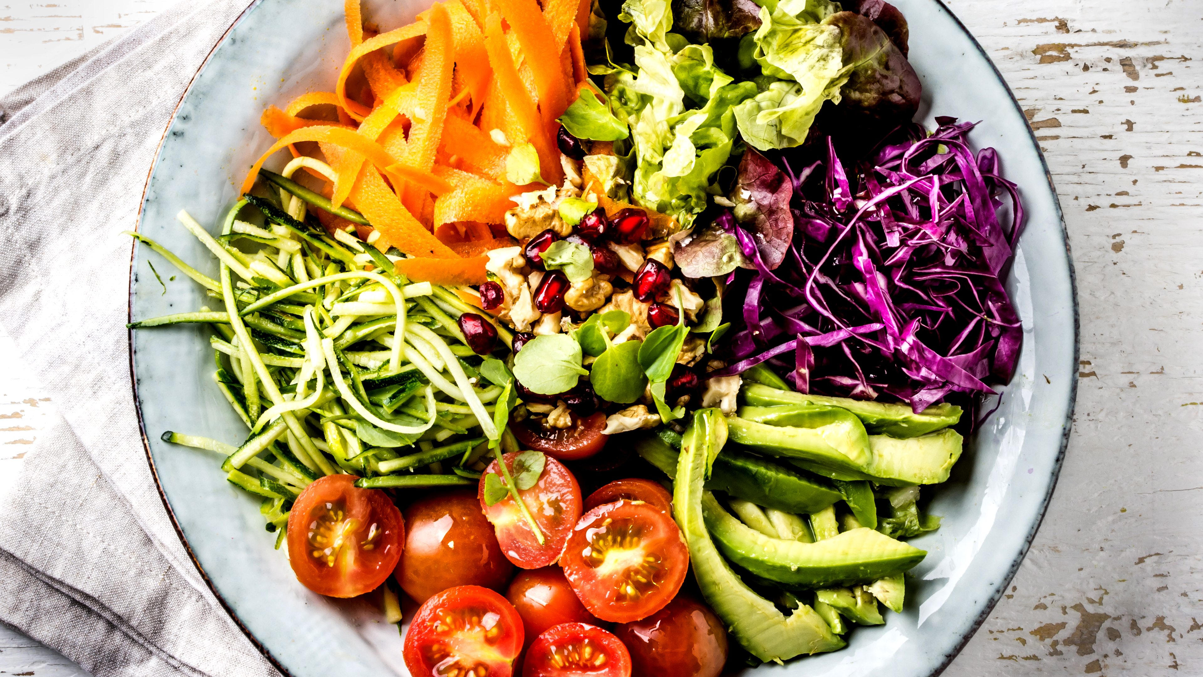 Vegan buddha bowl. Bowl with fresh raw vegetables - cabbage, carrot, zucchini, lettuce, watercress salad, tomatoes cherry and avocado, nuts and pomegranate on white background